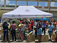 PuertoRico Five people in line getting Coleman gas stoves and gas canisters under a Ford CENTRO DE MOVILIDAD SOCIAL pop-up tent with numerous others and staff in the background near other tents.